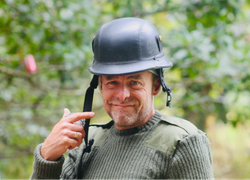 Man wearing a black helmet and green sweater, smiling and pointing at his helmet