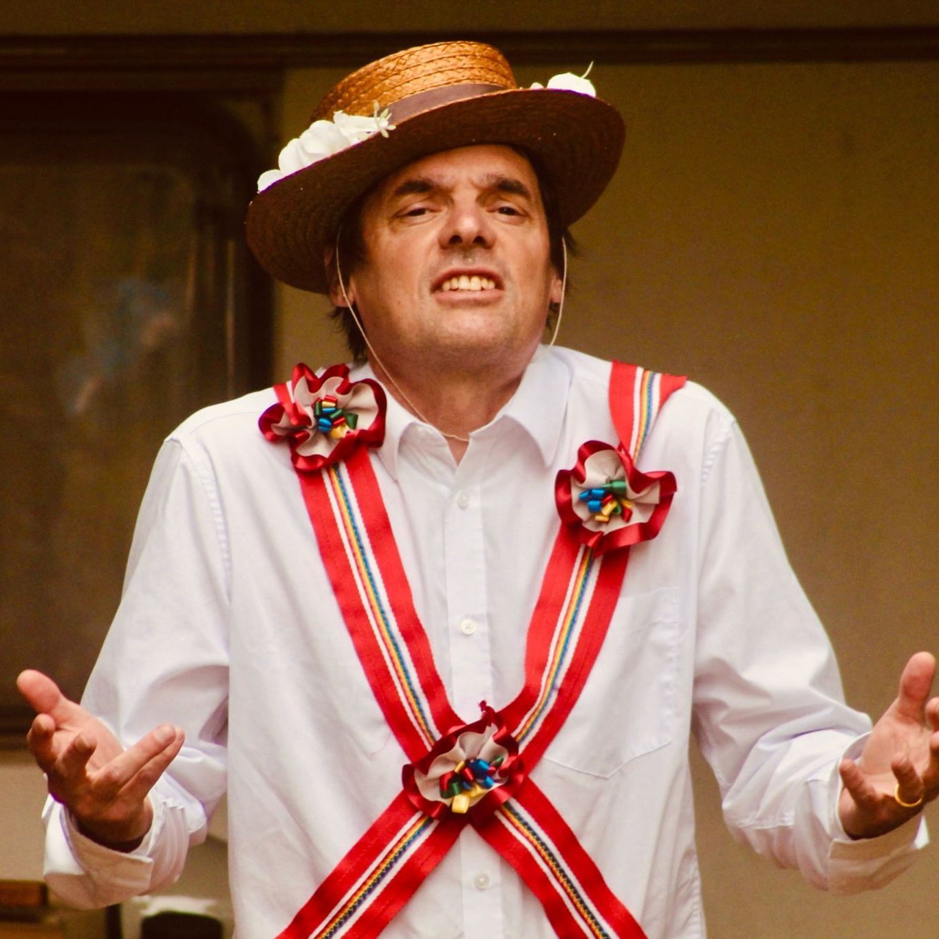 Man wearing a straw hat with flowers and a white shirt decorated with colorful ribbons and rosettes, gesturing with his hands.