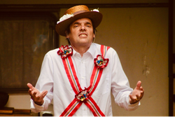 Man wearing a straw hat with flowers and a white shirt decorated with colorful ribbons and rosettes, gesturing with his hands.