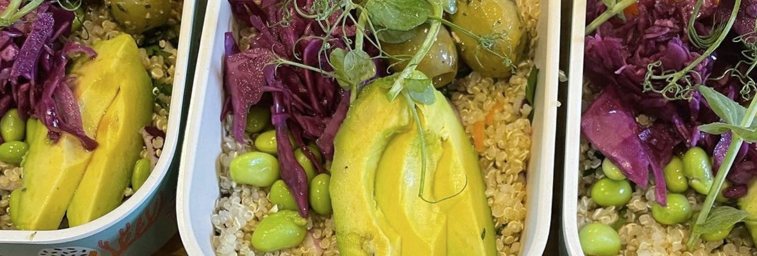 Containers filled with colorful quinoa bowls topped with avocado, roasted sweet potatoes, olives, edamame, red cabbage, and greens.