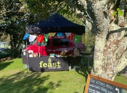 Outdoor food stand named Feast Kitchen Deli with a black canopy, colorful flags, and a chalkboard menu