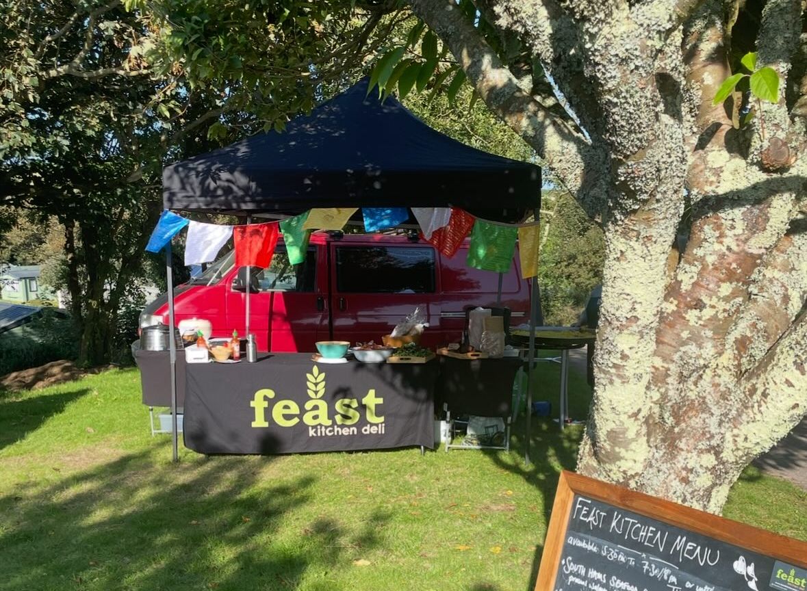 Outdoor food stand named Feast Kitchen Deli with a black canopy, colorful flags, and a chalkboard menu