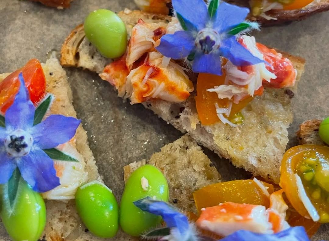 Crostini with yellow and orange cherry tomatoes, green edamame, shredded crab meat, and purple edible flowers