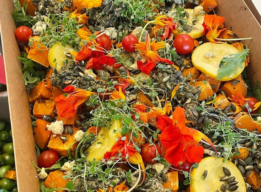 Colorful mixed vegetable salad with edible flowers, cherry tomatoes, squash, pumpkin cubes, seeds, leafy greens, and microgreens in a cardboard box.
