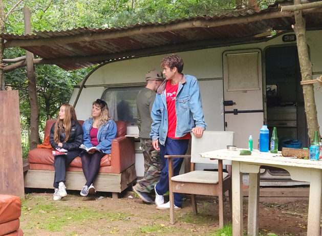 People gathered outside a caravan in a wooded area, sitting on mismatched furniture under a makeshift shelter.