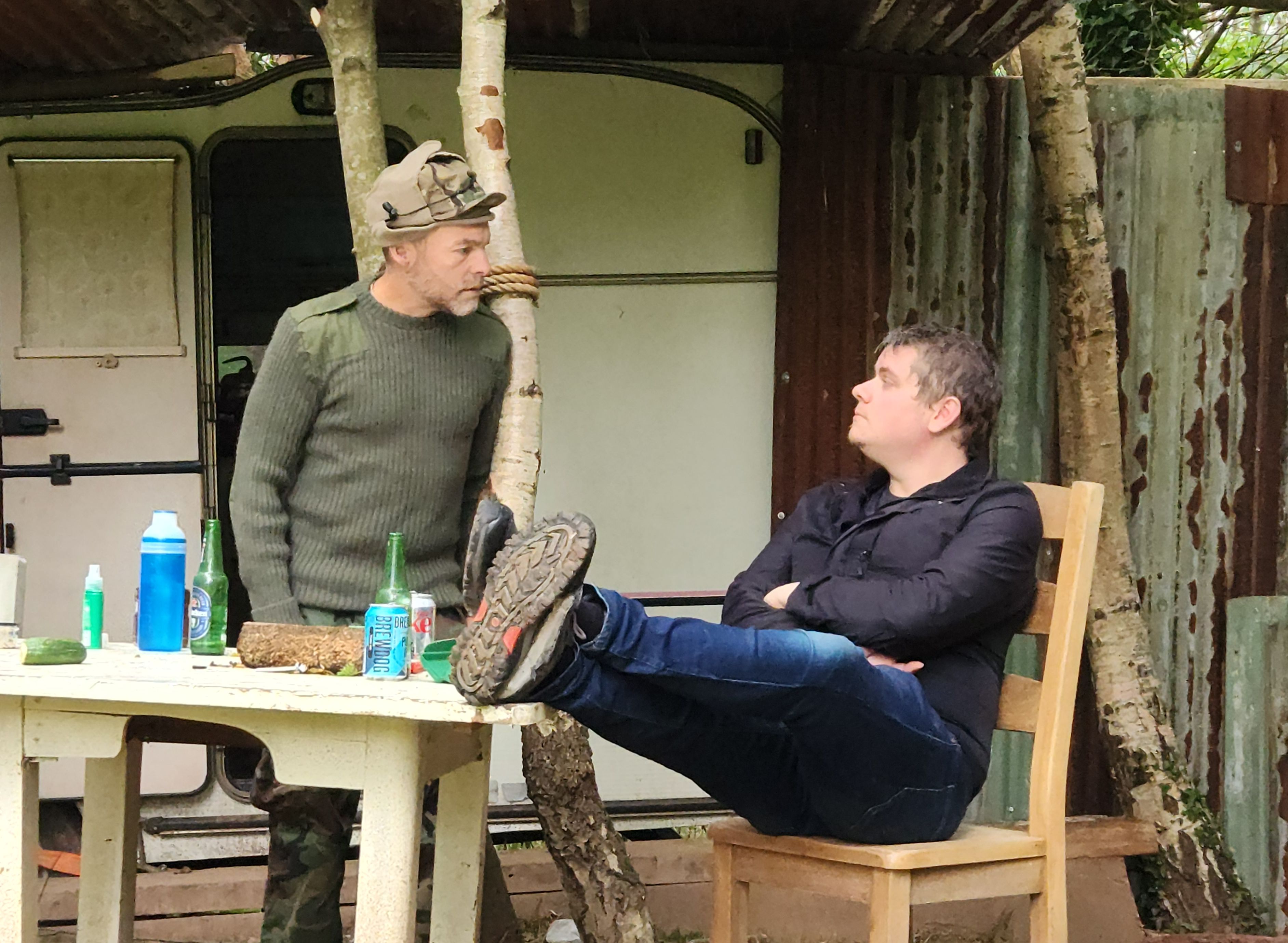 Two men talking at an outdoor table in a rustic, wooded setting with a shed and a caravan in the background.
