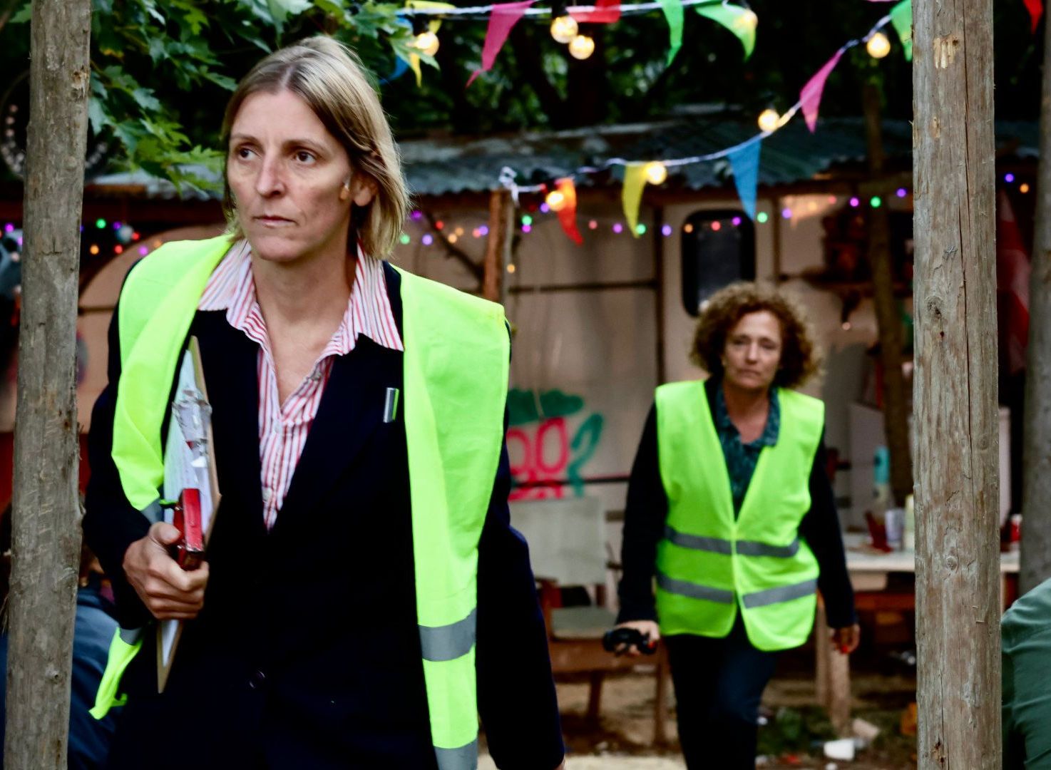 Two women wearing yellow reflective vests in an outdoor area decorated with colorful bunting and string lights.
