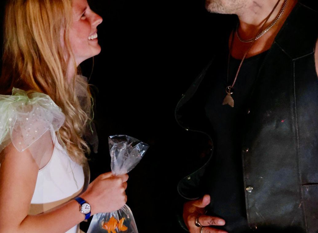 A woman in a light sparkly outfit holding a bag with a goldfish, smiling at a man with curly hair and a leather vest in dim lighting.
