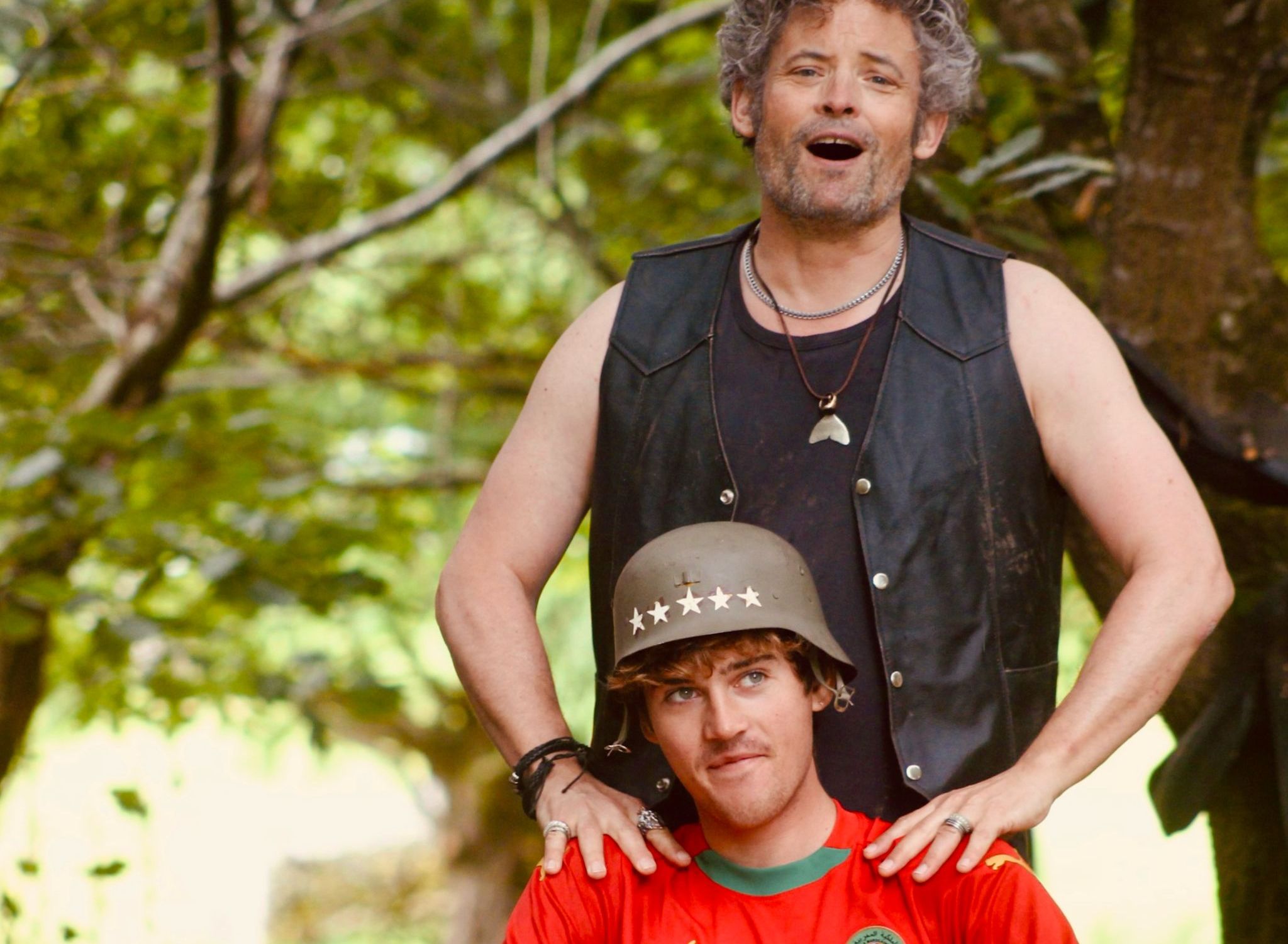 Two men, one standing and one sitting, outdoors. The standing man is wearing a sleeveless black leather vest and the seated man is wearing a helmet and a red sports jersey.