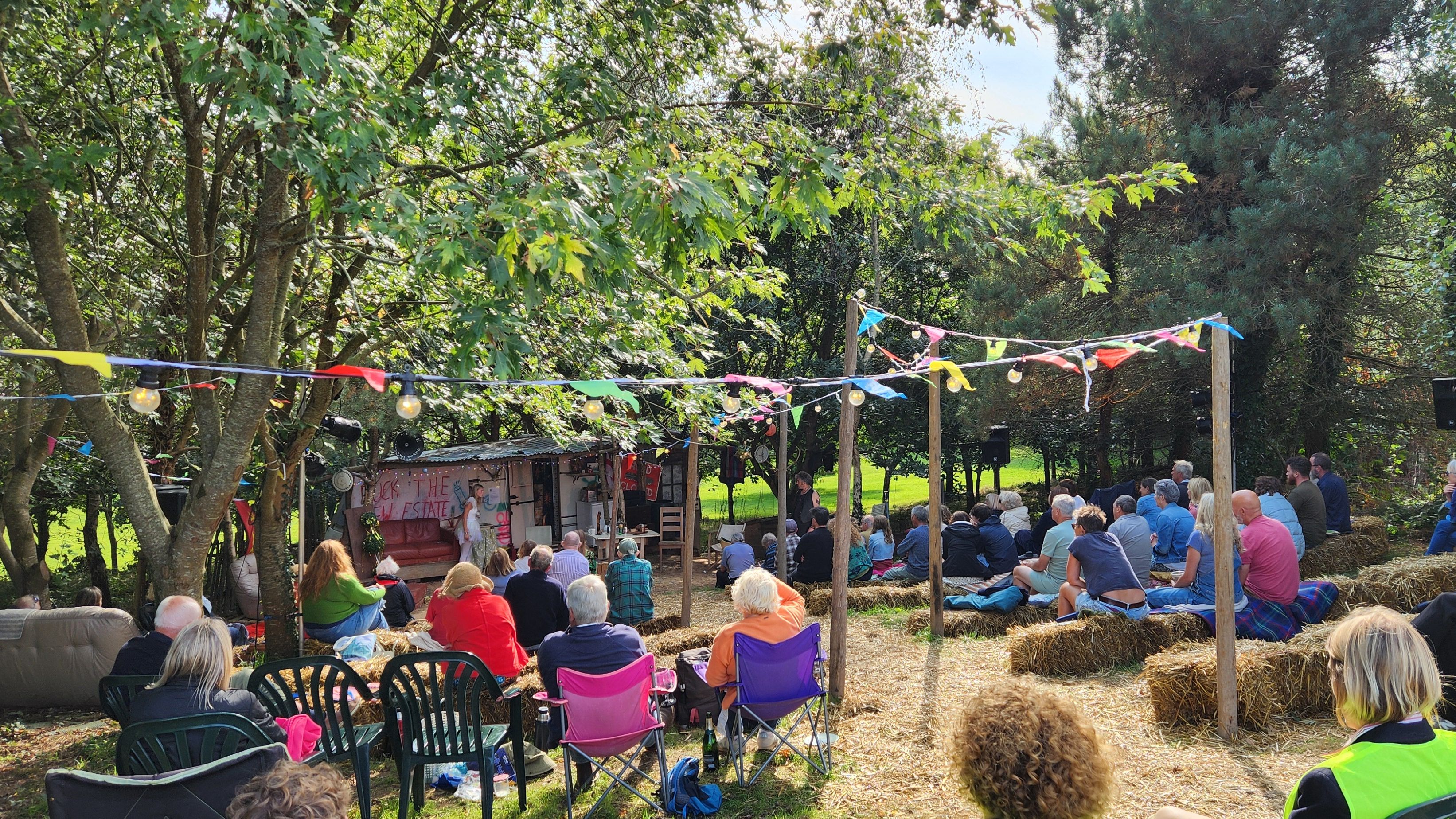 People sitting on hay bales and chairs at an outdoor event under colorful bunting in a wooded area.