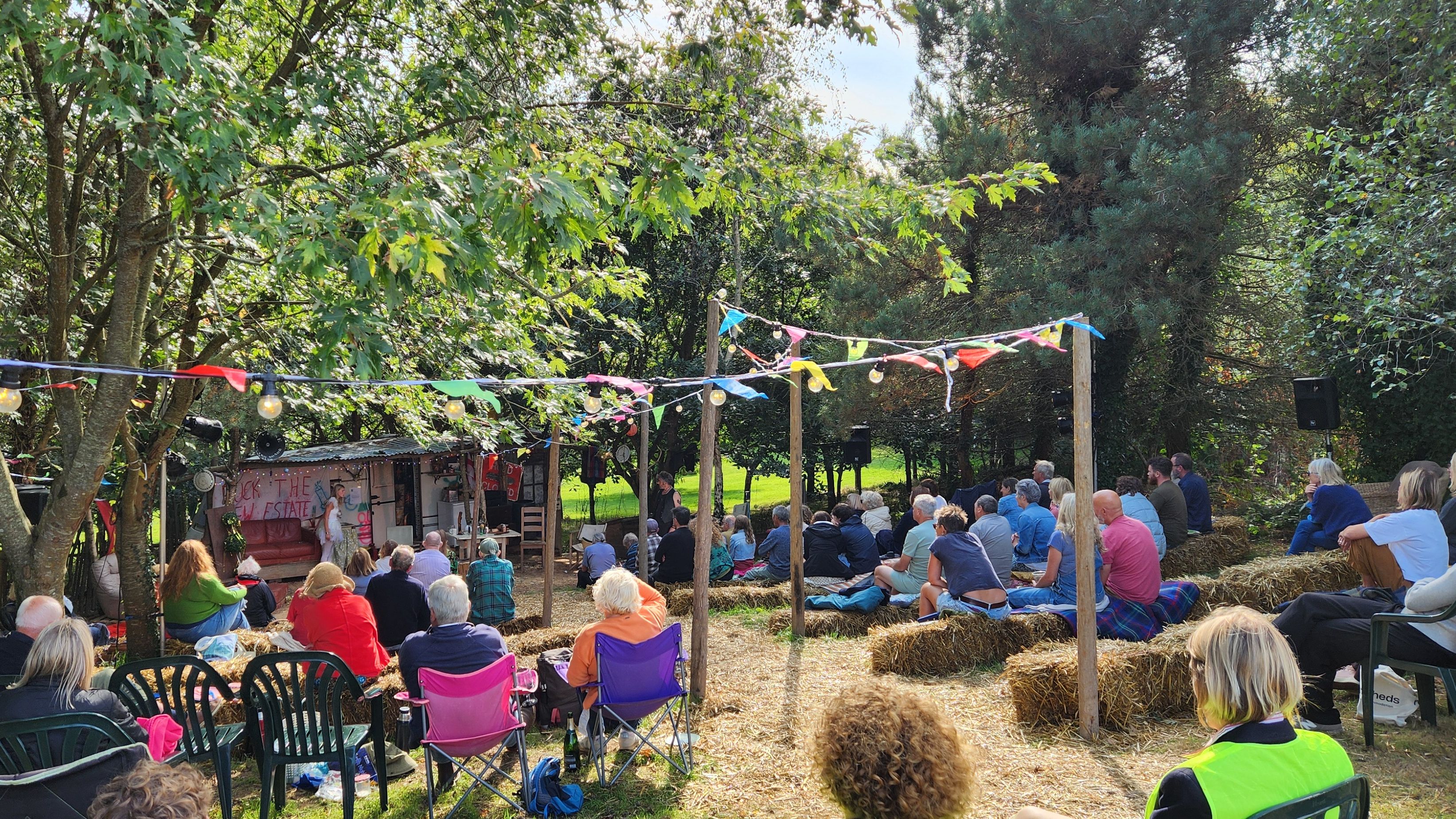 People sitting on hay bales and chairs at an outdoor event under colorful bunting in a wooded area.