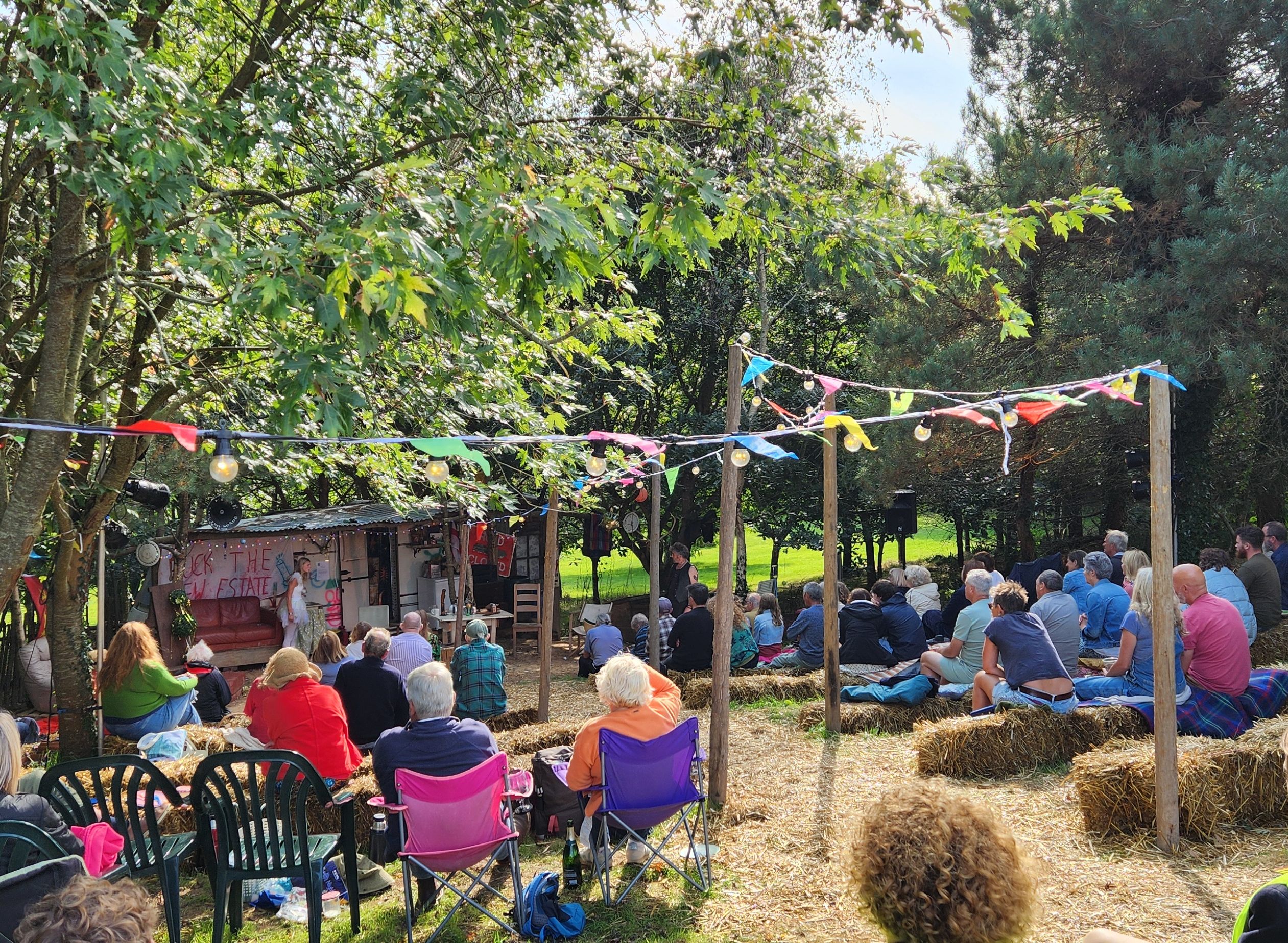 People sitting on hay bales and chairs at an outdoor event under colorful bunting in a wooded area.