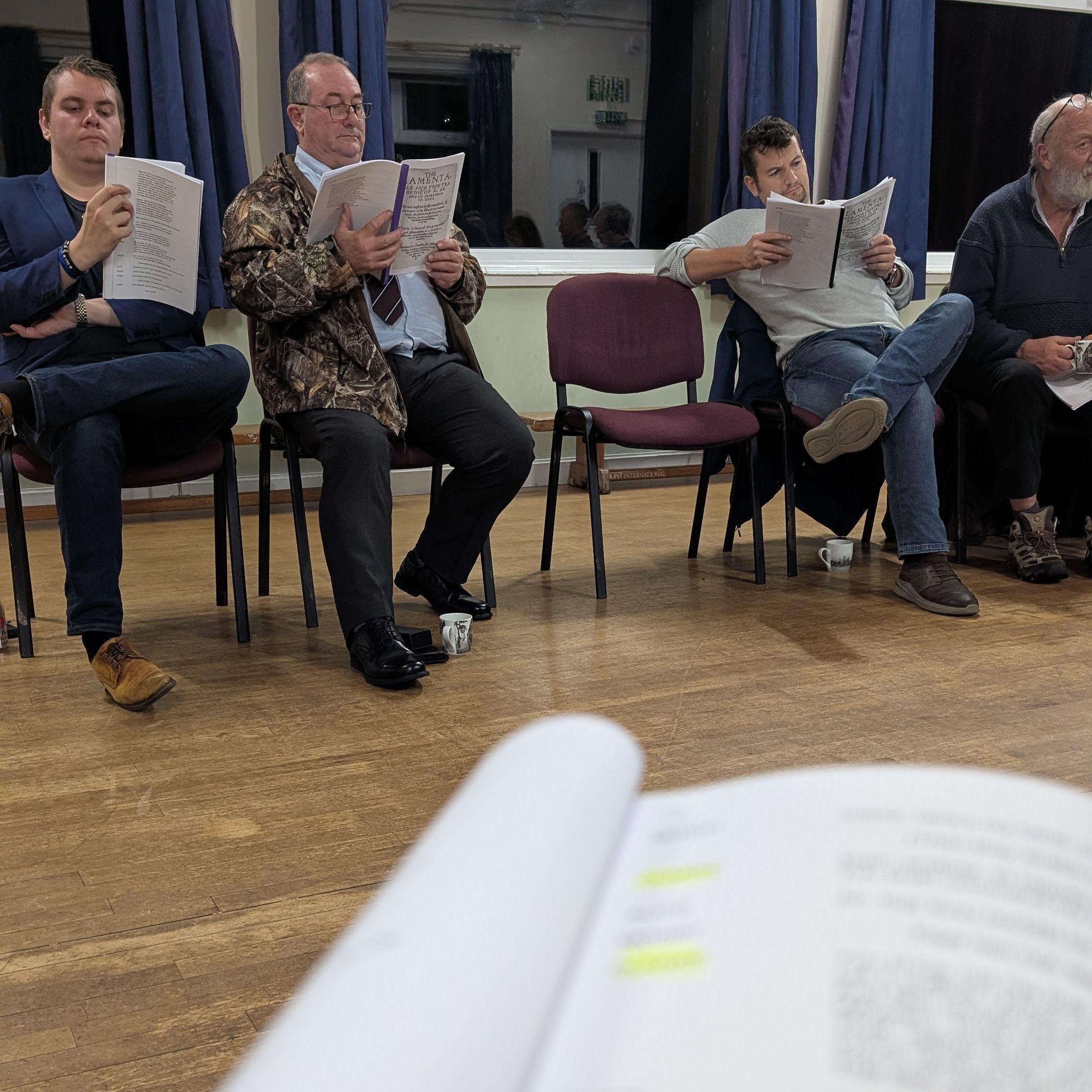 A group of people seated in a semicircle, each holding a script and reading, with an open script in the foreground out of focus.