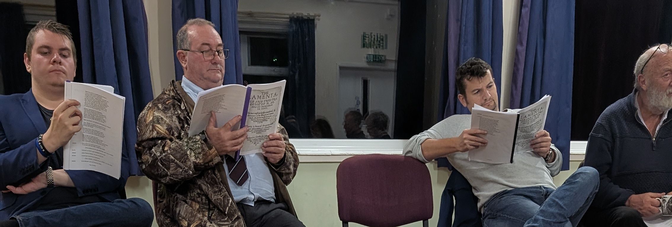A group of people seated in a semicircle, each holding a script and reading, with an open script in the foreground out of focus.