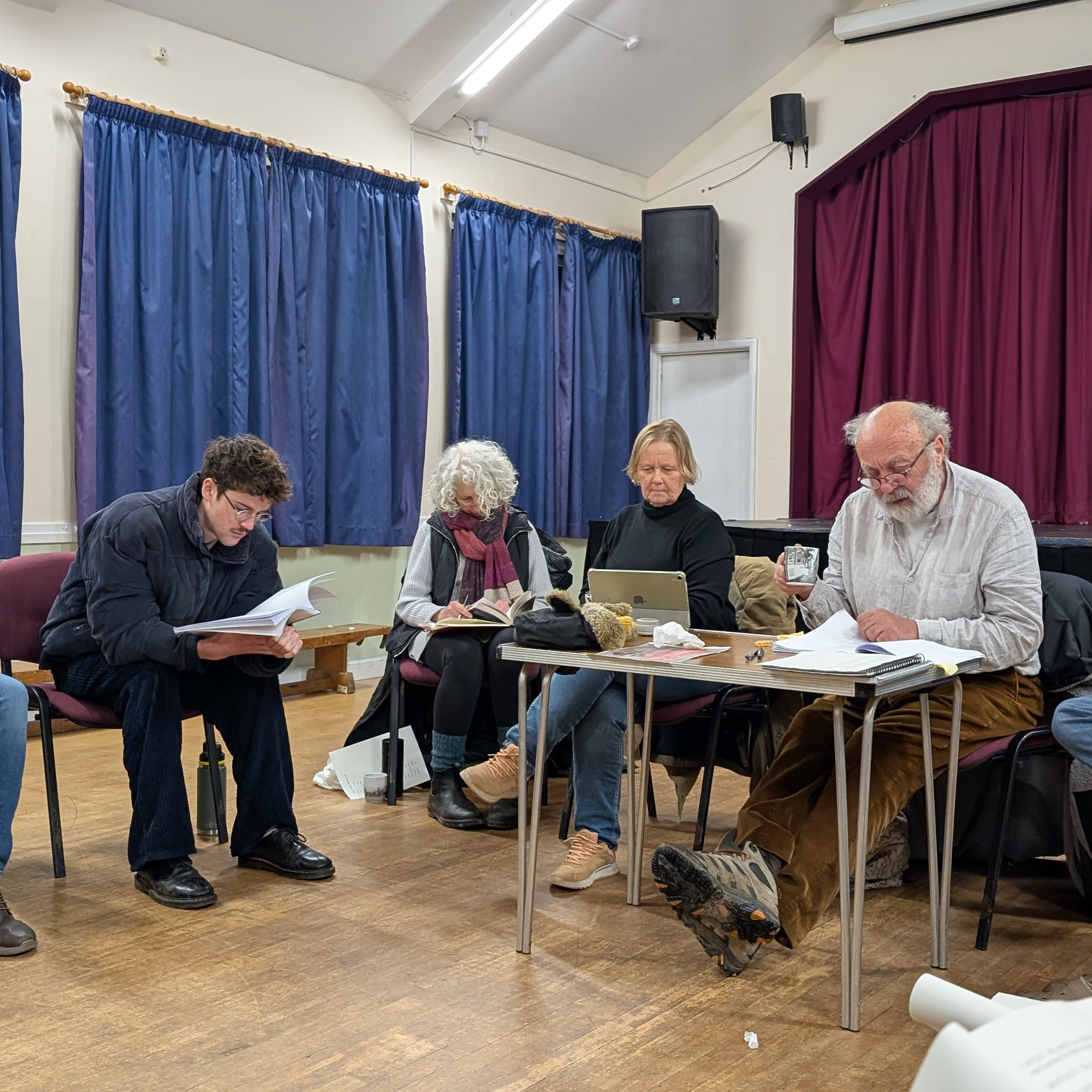 A group of people sitting in a circle reading scripts in a room with blue curtains and a small stage.
