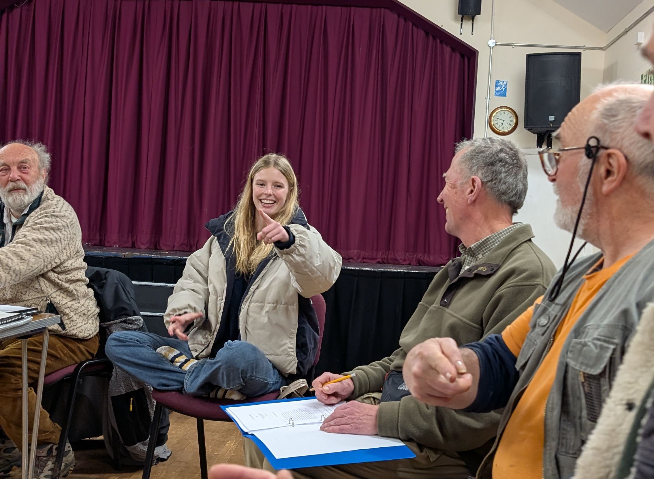 Group of people sitting in a room with a stage and maroon curtain in the background. One young woman is smiling and pointing, others are engaged in conversation.