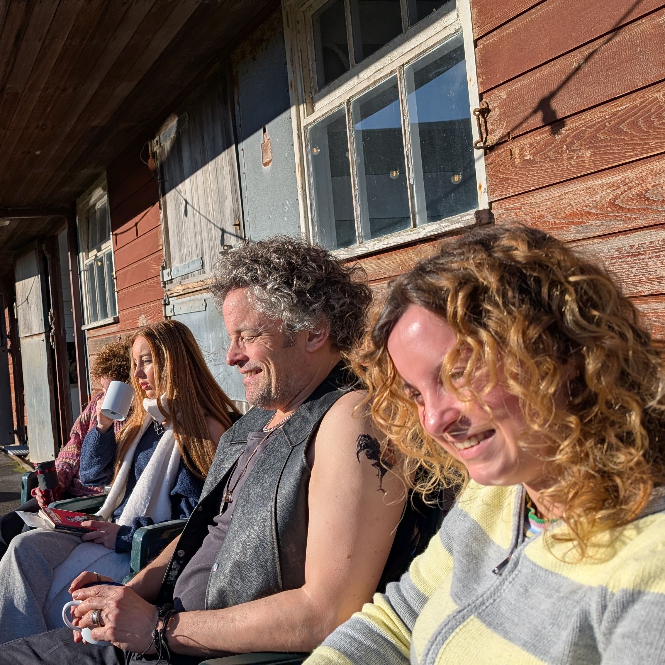 Group of people sitting outside on chairs next to a rustic wooden building, enjoying sunny weather.