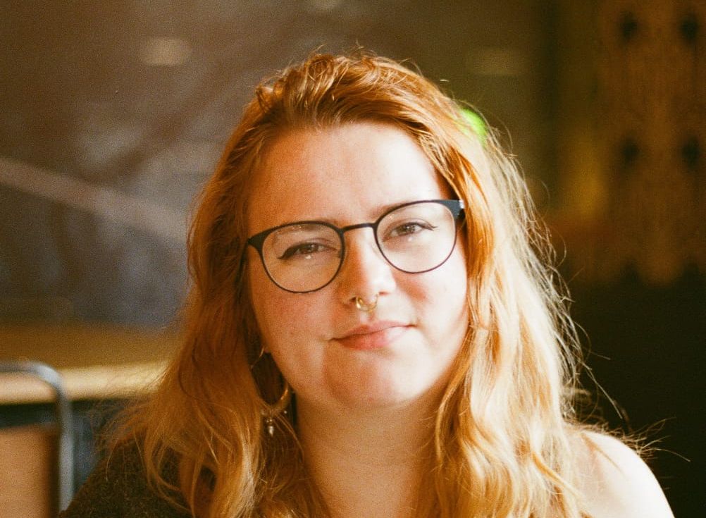 A woman with long wavy hair, glasses, and a nose ring sitting indoors.