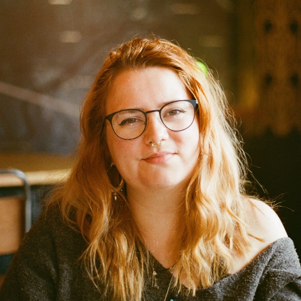 A woman with long wavy hair, glasses, and a nose ring sitting indoors.