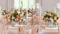 Baby pink, white and green flower arrangements on table with long candle sticks, and chairs with baby pink chair wraps