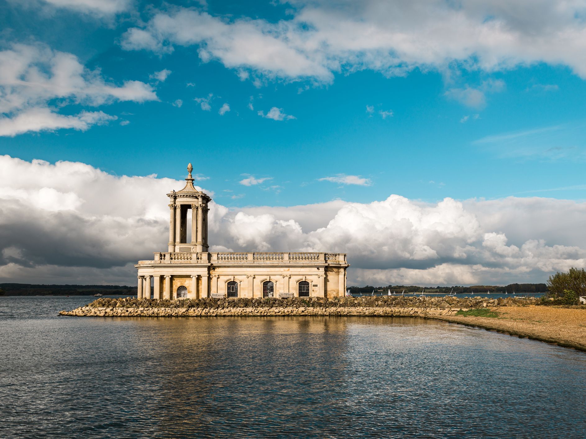 A historic stone church with columns and a domed tower stands partially submerged in a reservoir, surrounded by a rocky shoreline, under a bright blue sky with fluffy clouds.