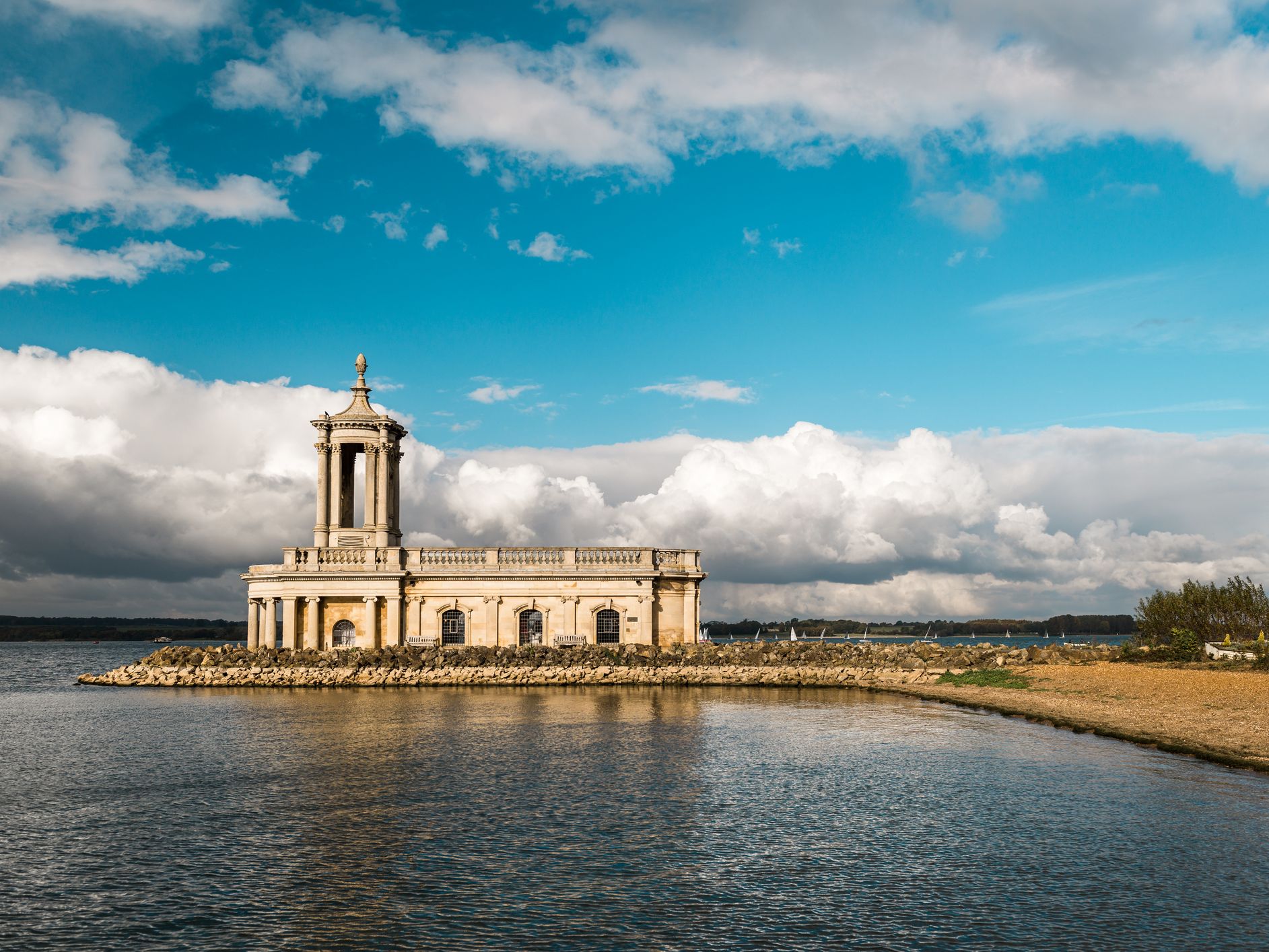 A historic stone church with columns and a domed tower stands partially submerged in a reservoir, surrounded by a rocky shoreline, under a bright blue sky with fluffy clouds.