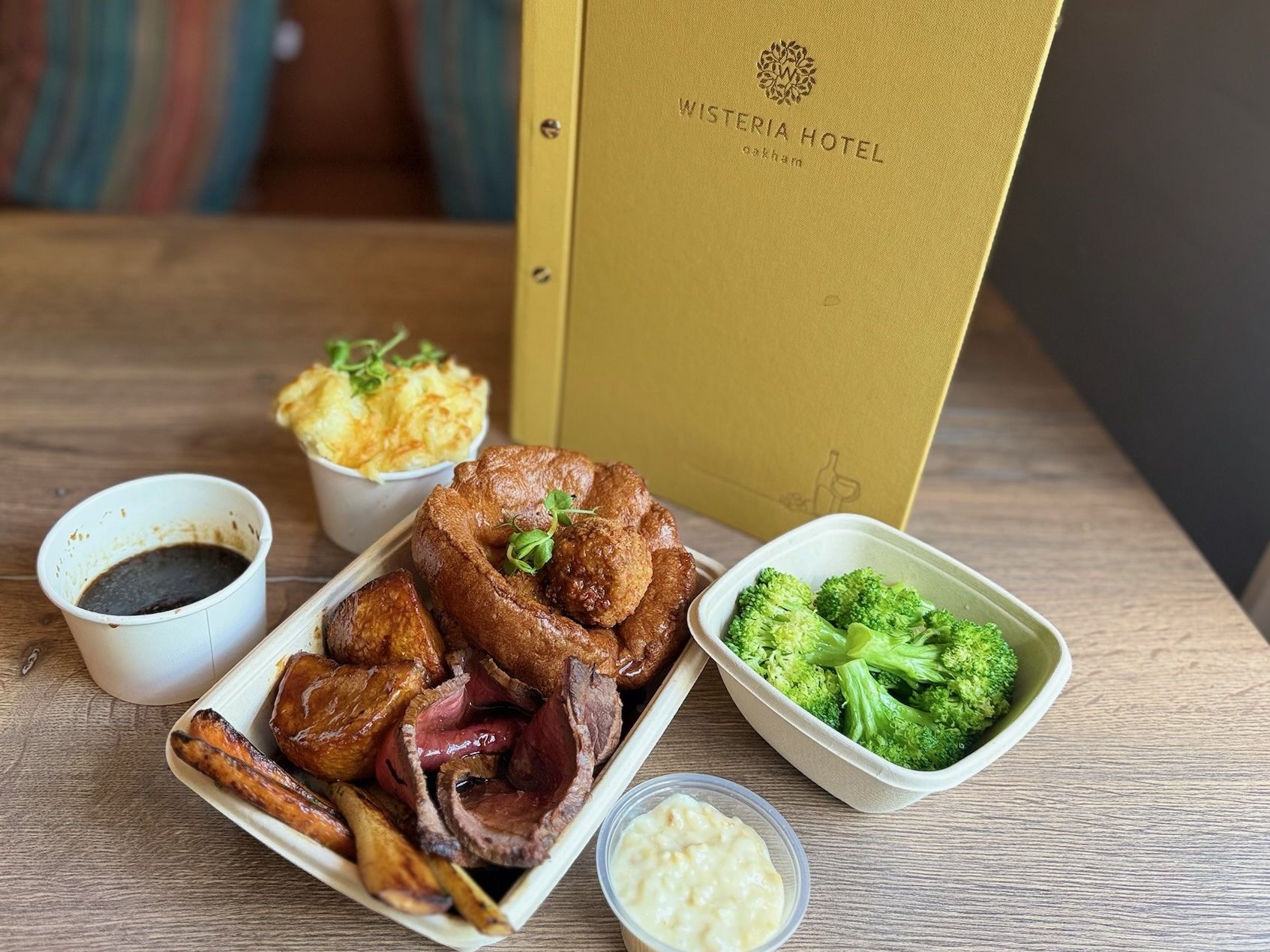 Traditional roast dinner with beef, vegetables, Yorkshire pudding, gravy, and sides, next to Wisteria Hotel menu on a table.