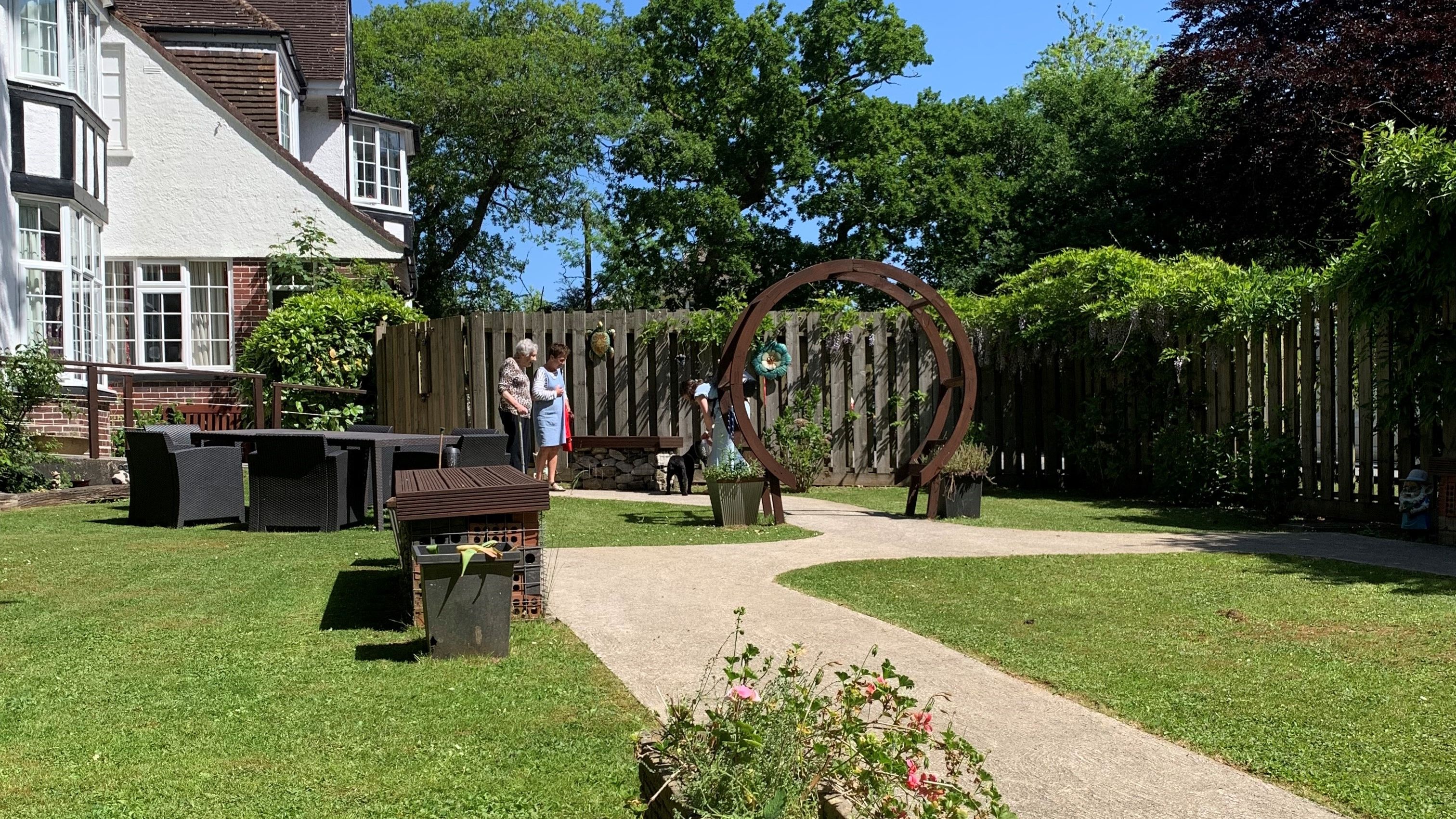 Sunny garden with a curved path, flower planter, outdoor seating, and a circular sculpture, with people chatting near a wooden fence and trees.