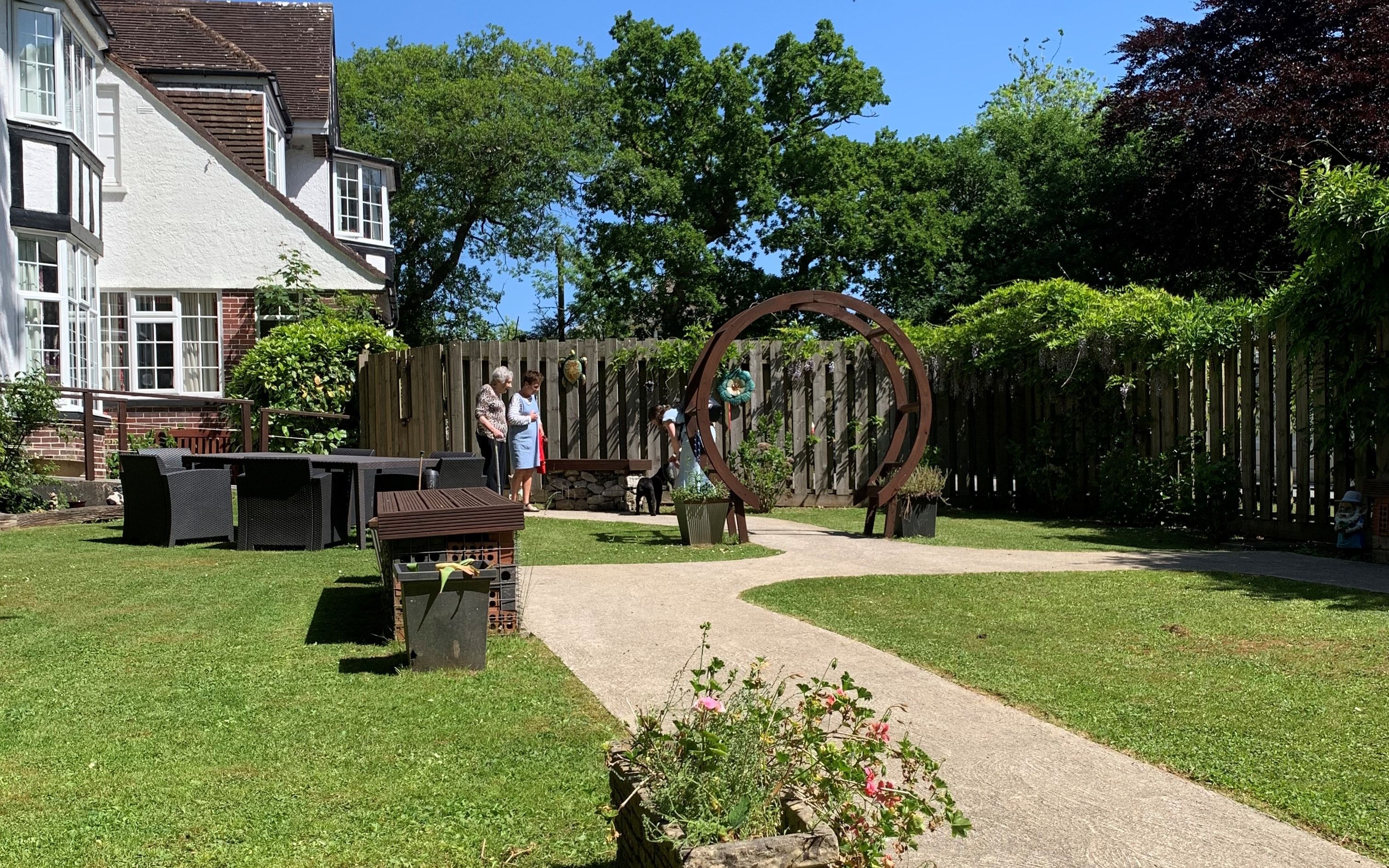Sunny garden with a curved path, flower planter, outdoor seating, and a circular sculpture, with people chatting near a wooden fence and trees.