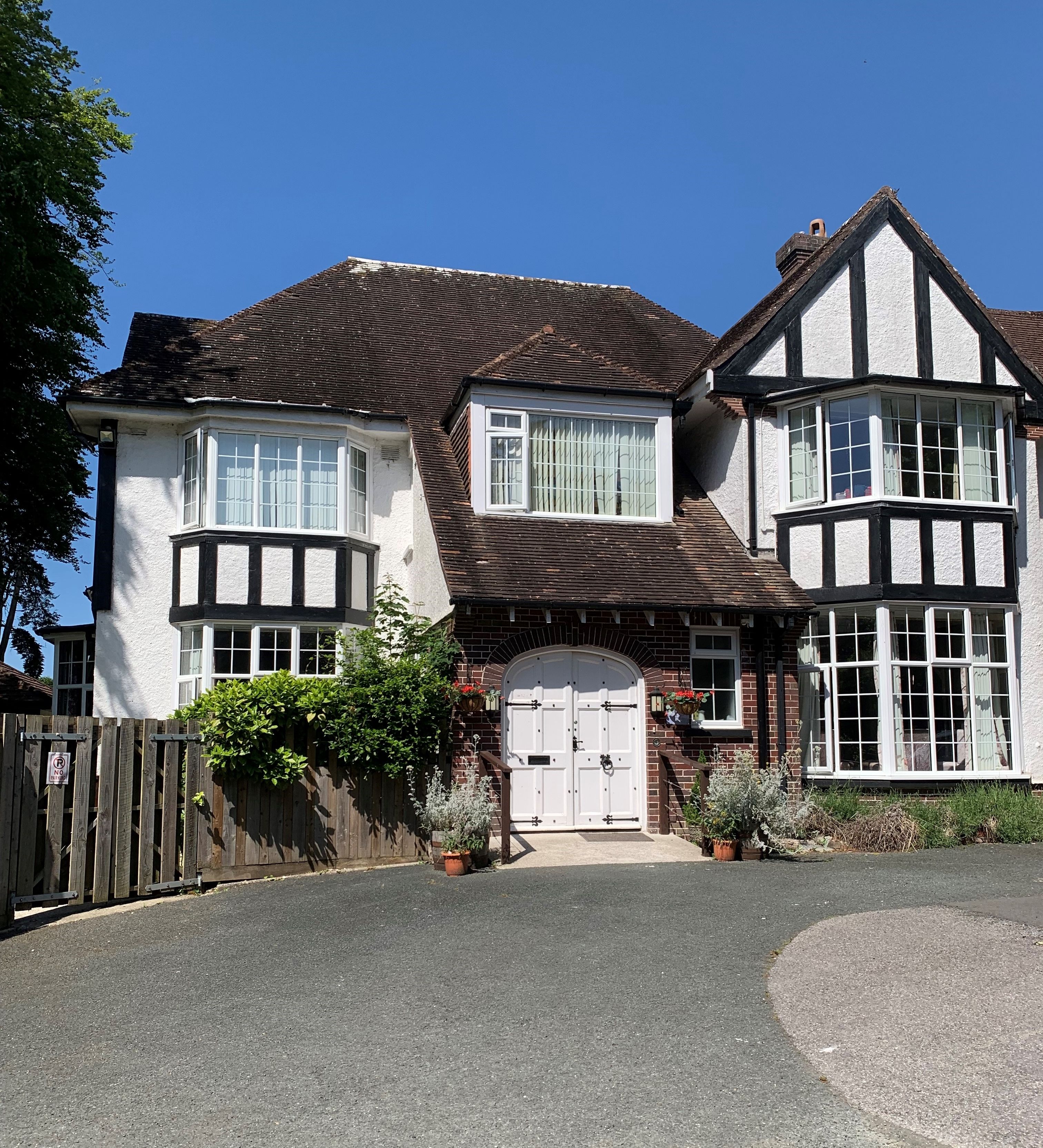 Tudor-style house with white and brick exterior, large bay windows, and a wide arched front door framed by potted plants and hanging baskets.