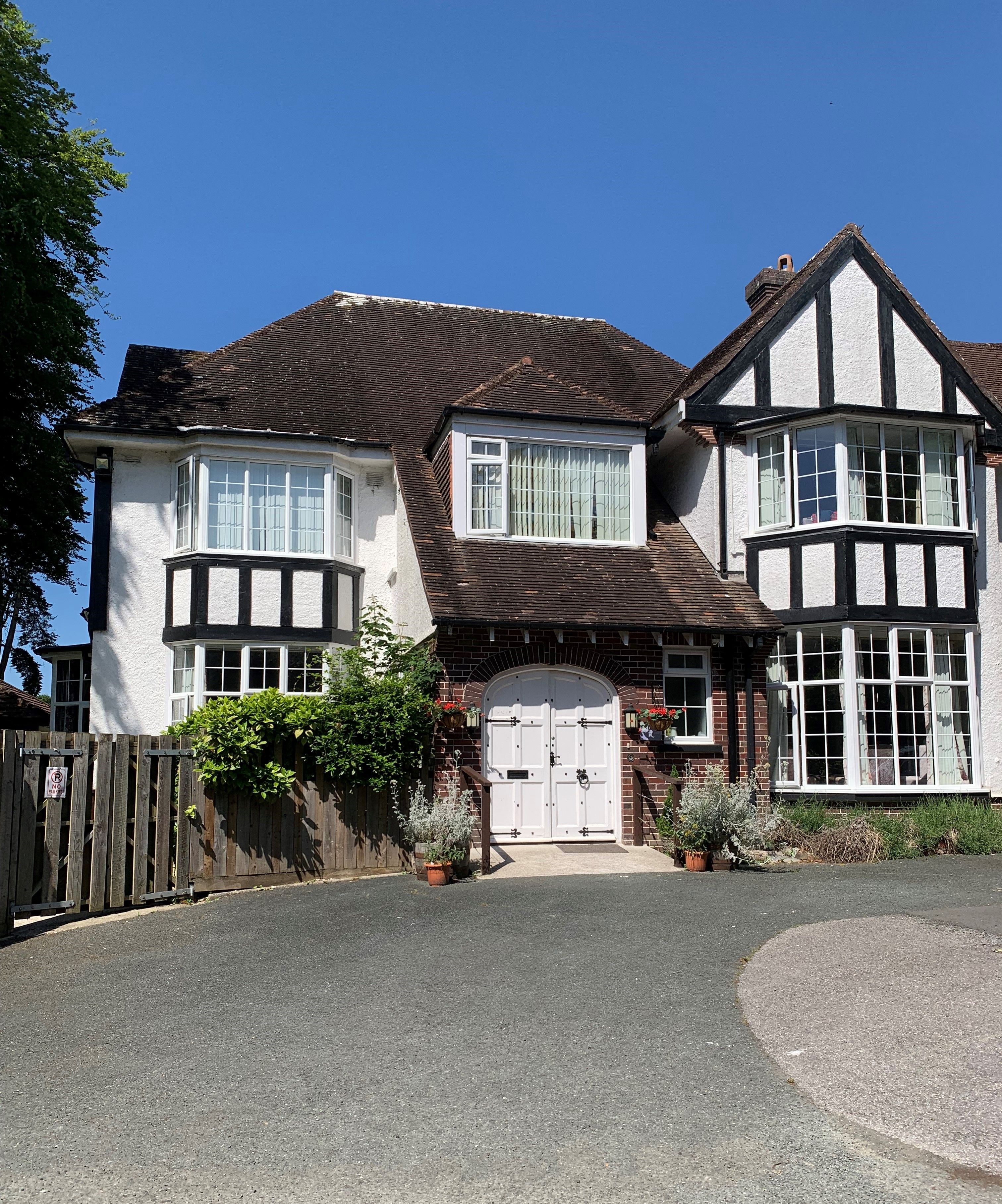 Tudor-style house with white and brick exterior, large bay windows, and a wide arched front door framed by potted plants and hanging baskets.