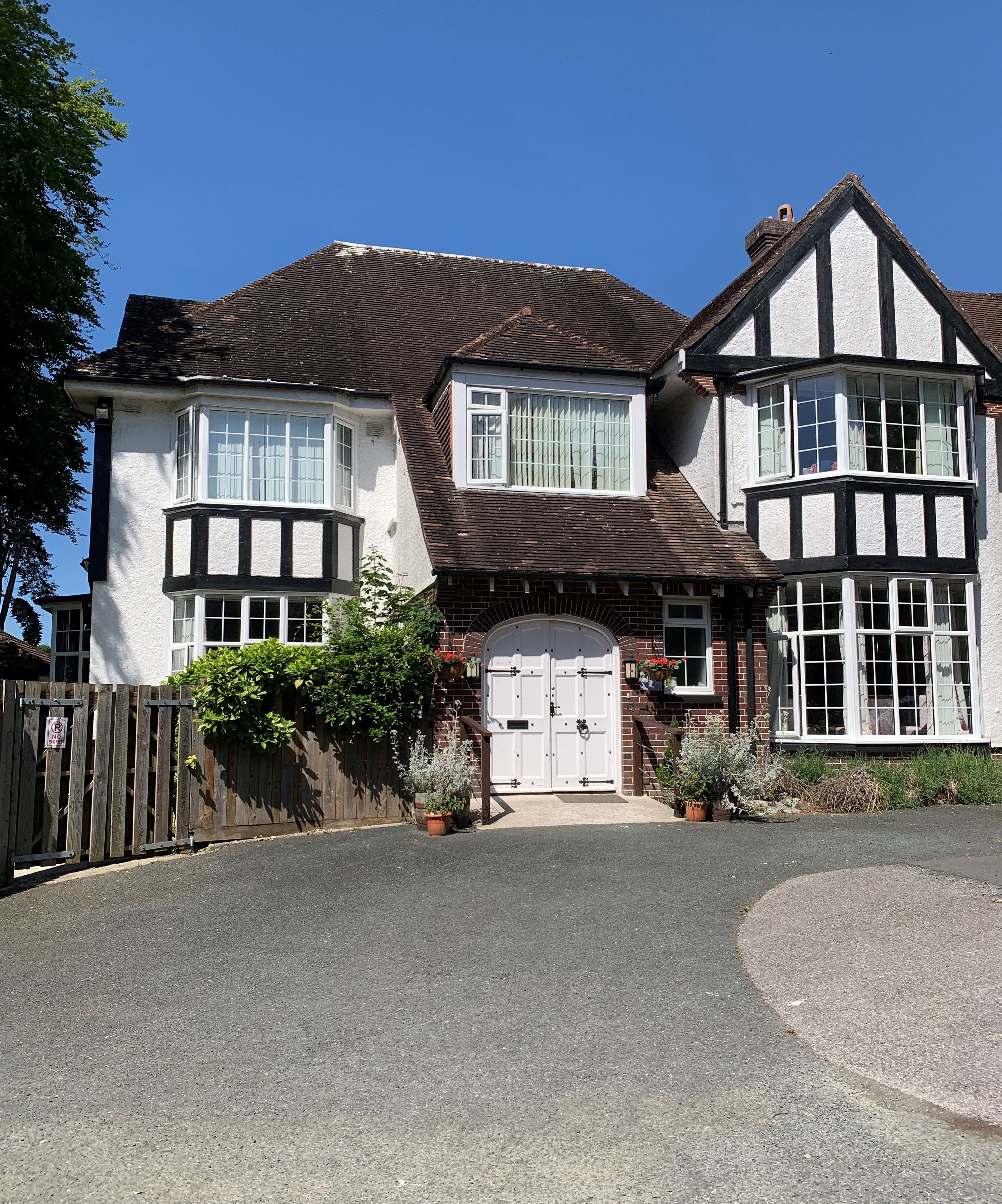 Tudor-style house with white and brick exterior, large bay windows, and a wide arched front door framed by potted plants and hanging baskets.