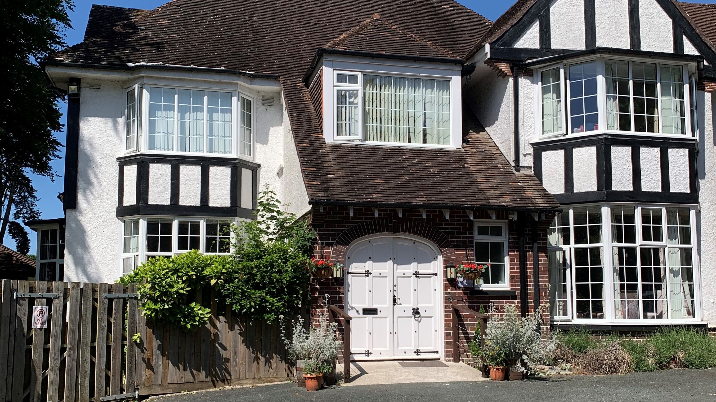 Tudor-style house with white and brick exterior, large bay windows, and a wide arched front door framed by potted plants and hanging baskets.