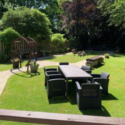 Outdoor dining set on lawn near a curved path and circular arch, surrounded by trees, plants, and a peaceful garden atmosphere.