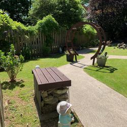 Curved garden path with a circular wooden arch, lush greenery, and a small statue in a sun hat holding flowers in the foreground.