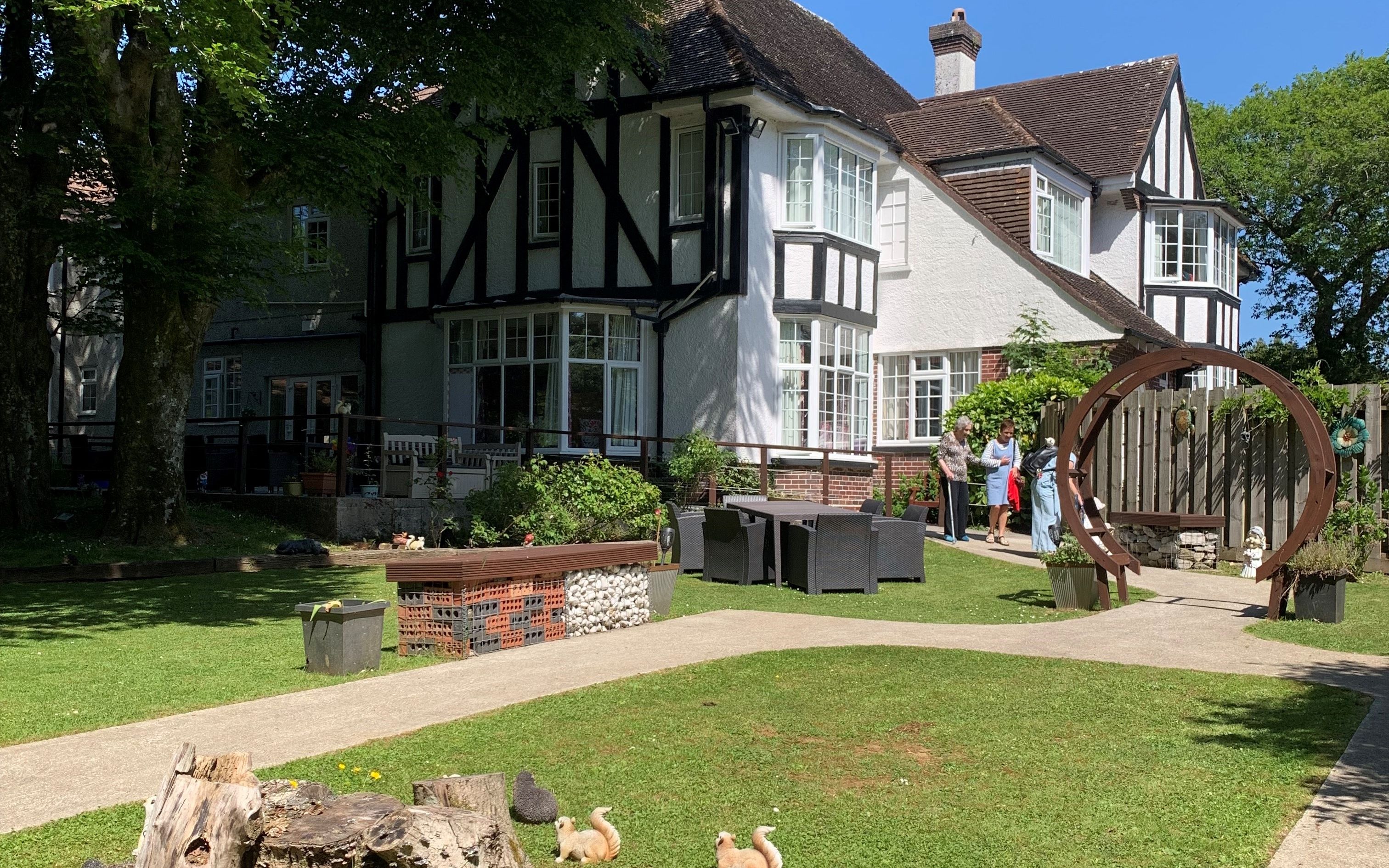Tudor-style house with a sunny garden, tree stump, animal statues, pink flowers, and a circular arch with people chatting nearby.