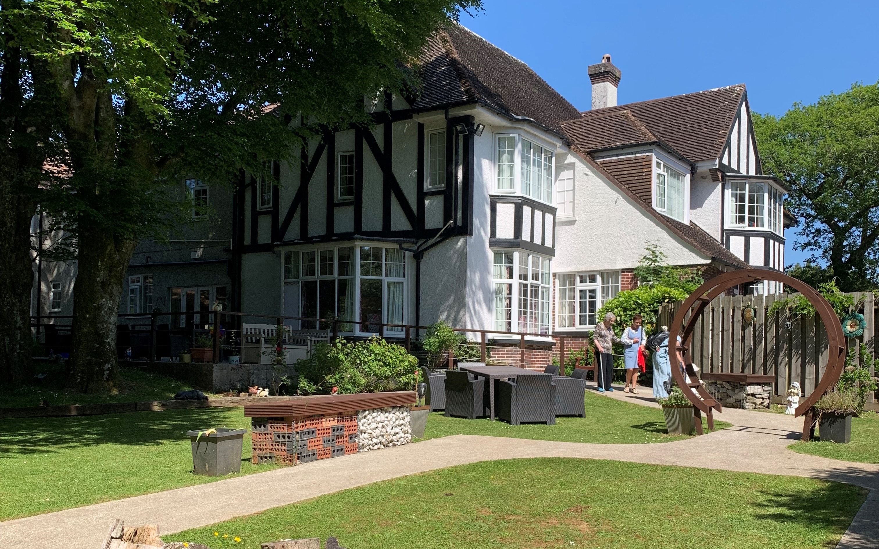 Tudor-style house with a sunny garden, tree stump, animal statues, pink flowers, and a circular arch with people chatting nearby.