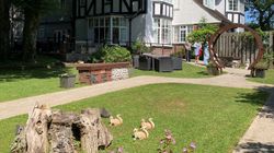Tudor-style house with a sunny garden, tree stump, animal statues, pink flowers, and a circular arch with people chatting nearby.