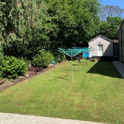 Side garden with rotary clotheslines, freshly washed laundry, a trimmed lawn, and dense trees lining one side of the path.