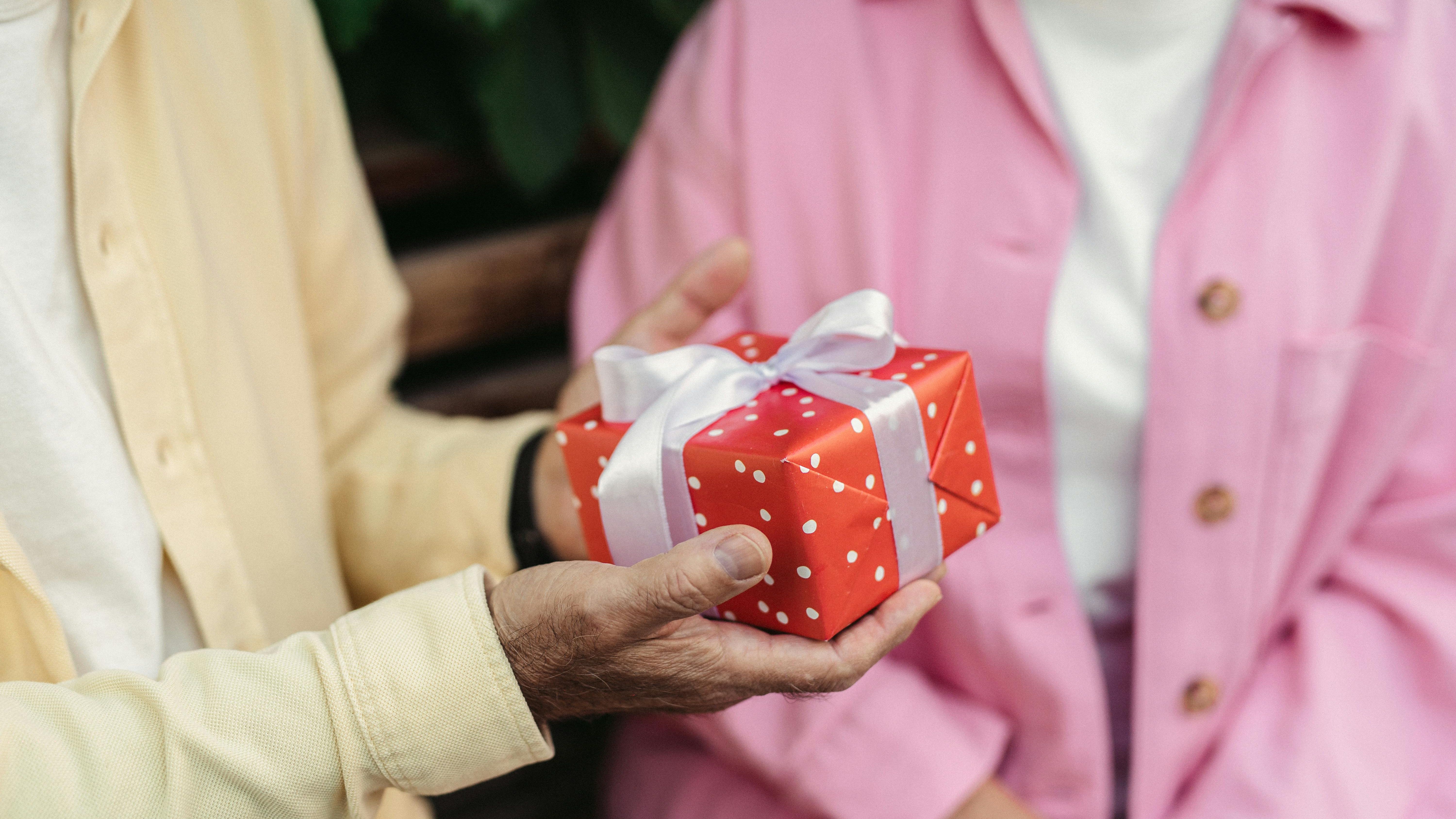 Close-up of a person in a yellow shirt handing a red gift box with white polka dots and a white ribbon to someone in a pink jacket.