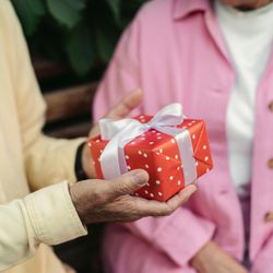 Close-up of a person in a yellow shirt handing a red gift box with white polka dots and a white ribbon to someone in a pink jacket.