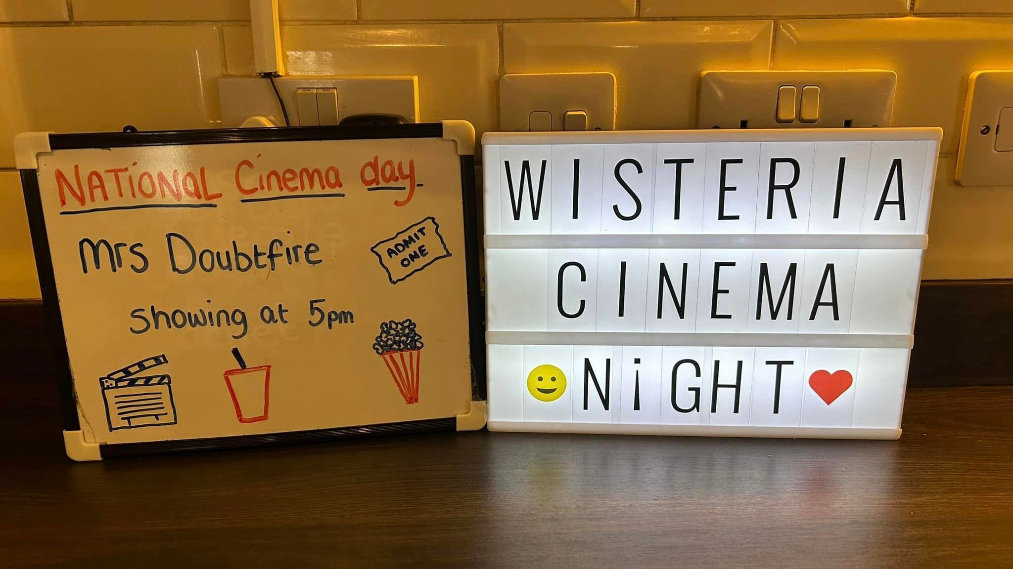 Whiteboard and lightbox on a counter announcing National Cinema Day, showing Mrs Doubtfire at 5 pm, with drawings of film, drink, popcorn, and emojis.