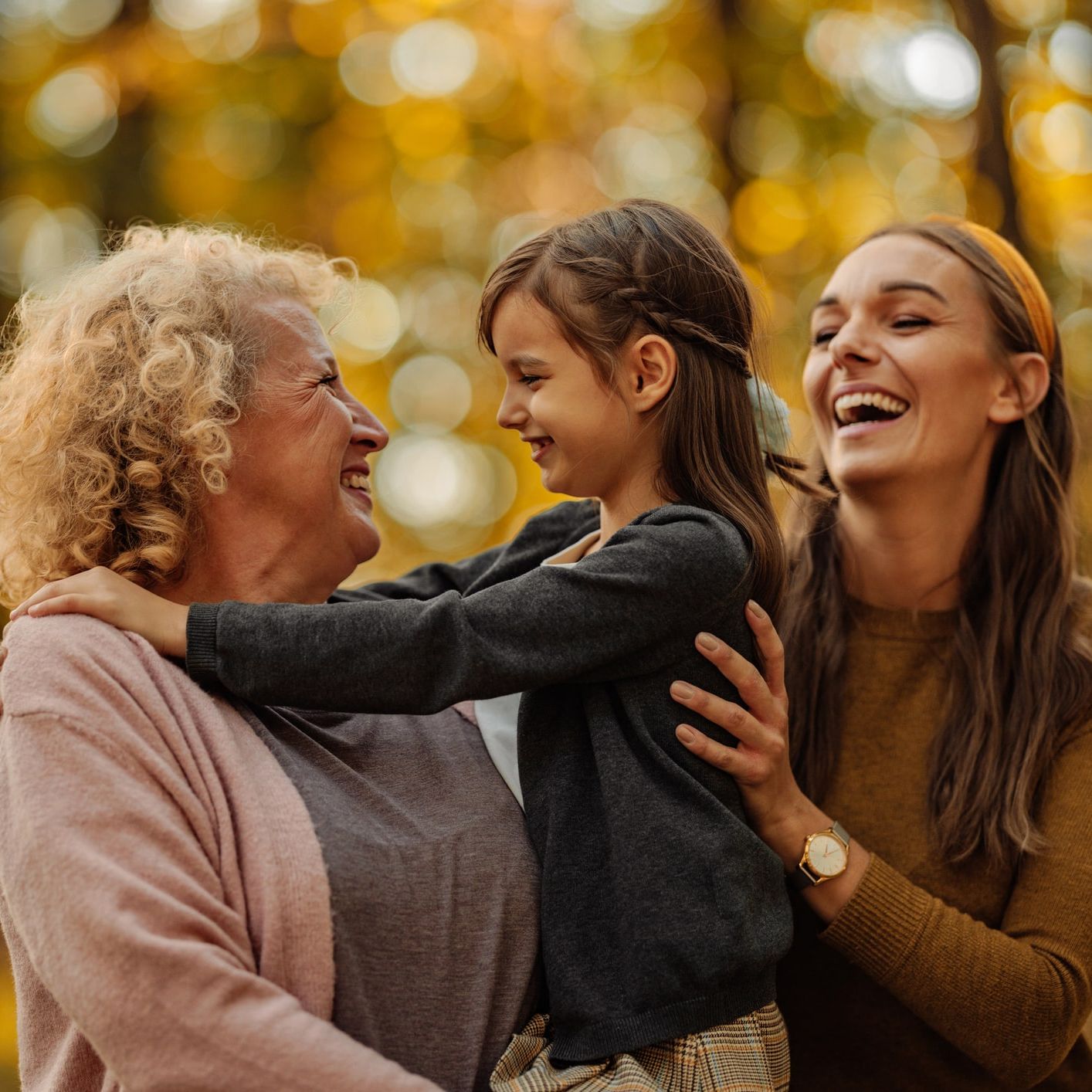 Three generations of women—grandmother, mother, and daughter—sharing a joyful moment together in a sunlit autumn forest.