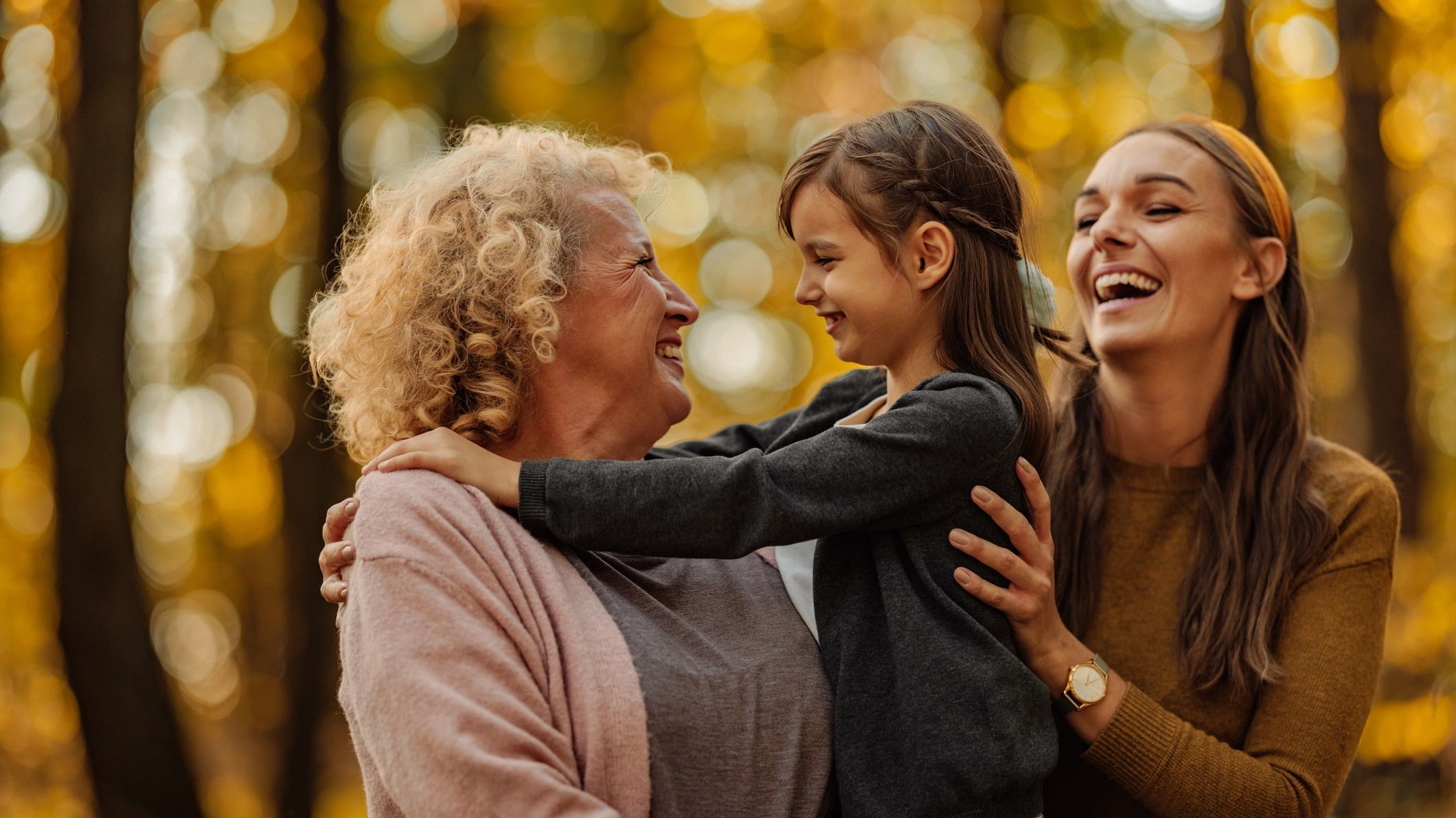 Three generations of women—grandmother, mother, and daughter—sharing a joyful moment together in a sunlit autumn forest.