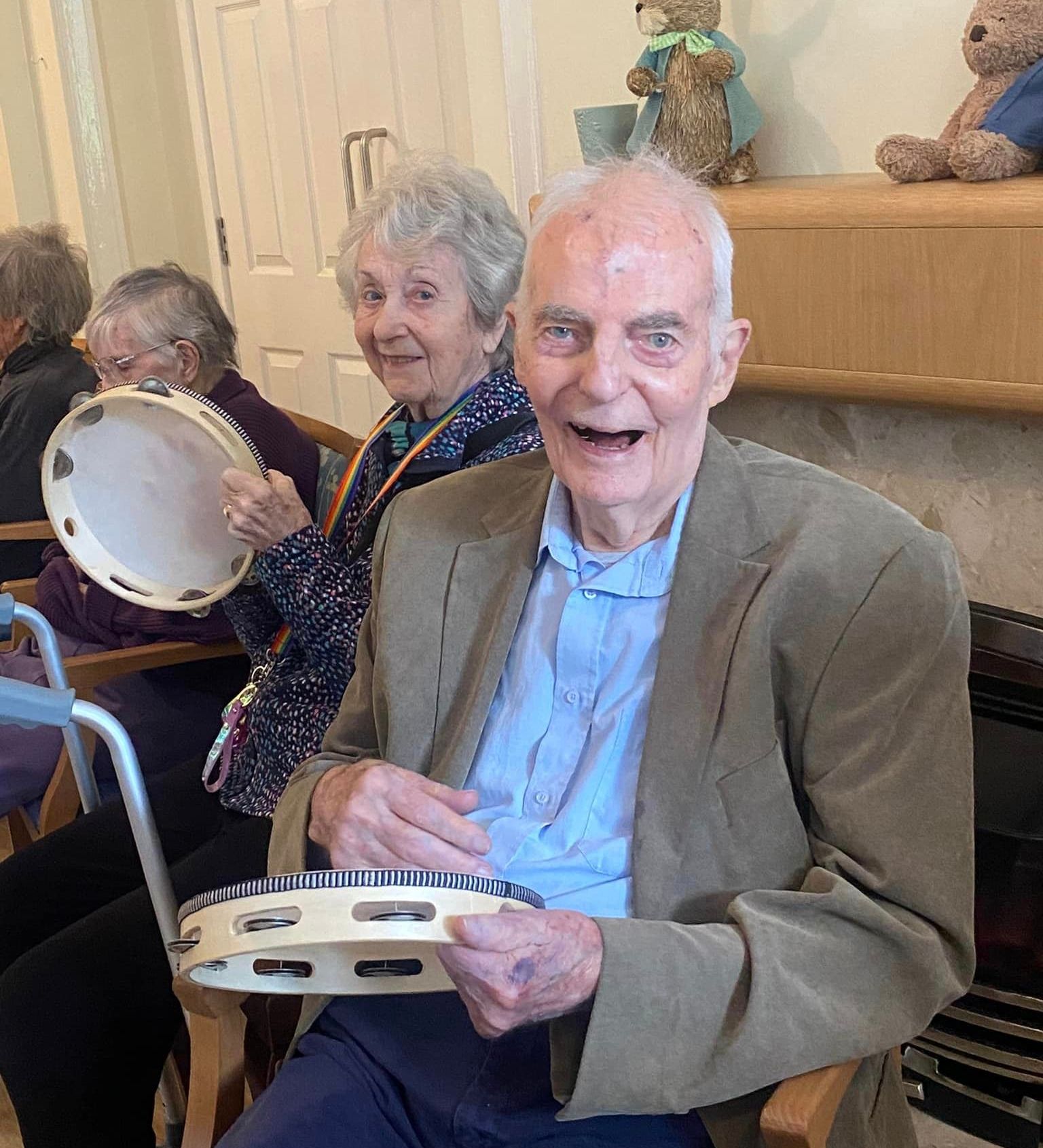 Elderly residents at Wisteria House Woolwell smiling and playing tambourines during a group activity, seated near a fireplace with plush toys above.
