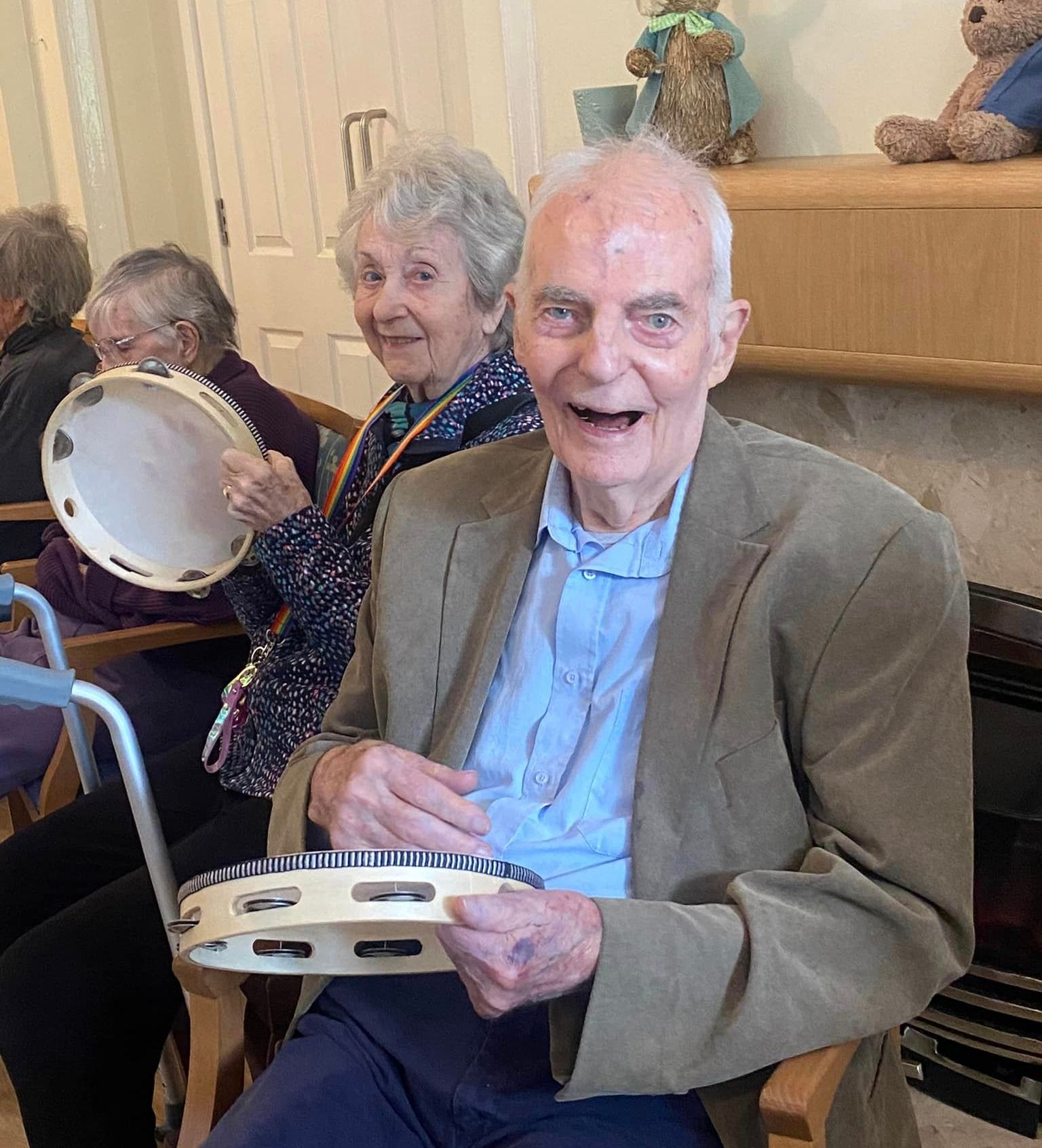 Elderly residents at Wisteria House Woolwell smiling and playing tambourines during a group activity, seated near a fireplace with plush toys above.