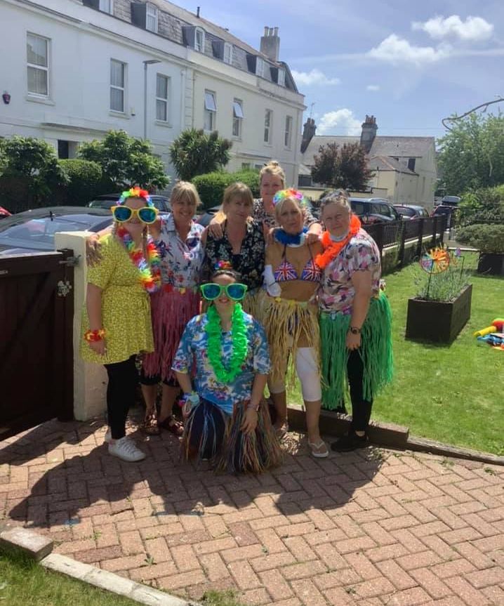 Staff at Wisteria Stoke dressed in colourful Hawaiian outfits and leis, smiling outdoors during a sunny party, with houses and greenery in the background.
