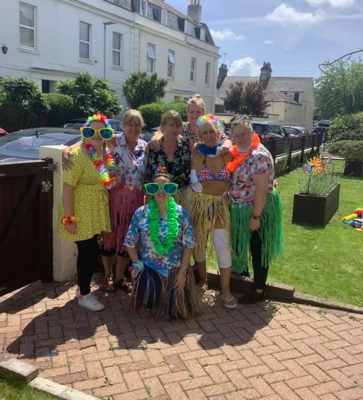 Staff at Wisteria Stoke dressed in colourful Hawaiian outfits and leis, smiling outdoors during a sunny party, with houses and greenery in the background.