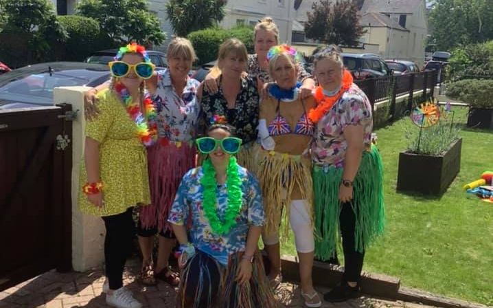 Staff at Wisteria Stoke dressed in colourful Hawaiian outfits and leis, smiling outdoors during a sunny party, with houses and greenery in the background.