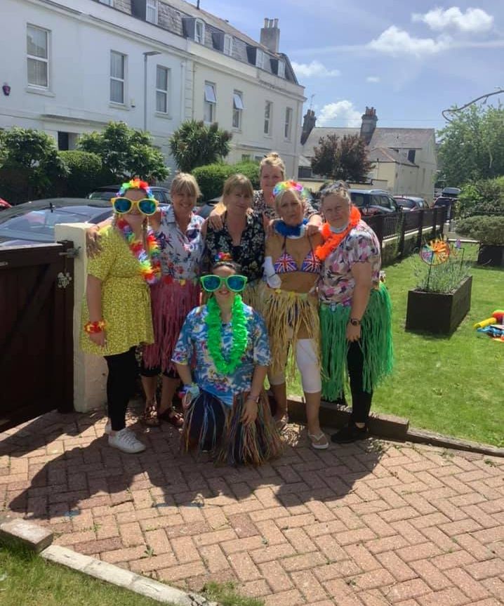 Staff at Wisteria Stoke dressed in colourful Hawaiian outfits and leis, smiling outdoors during a sunny party, with houses and greenery in the background.